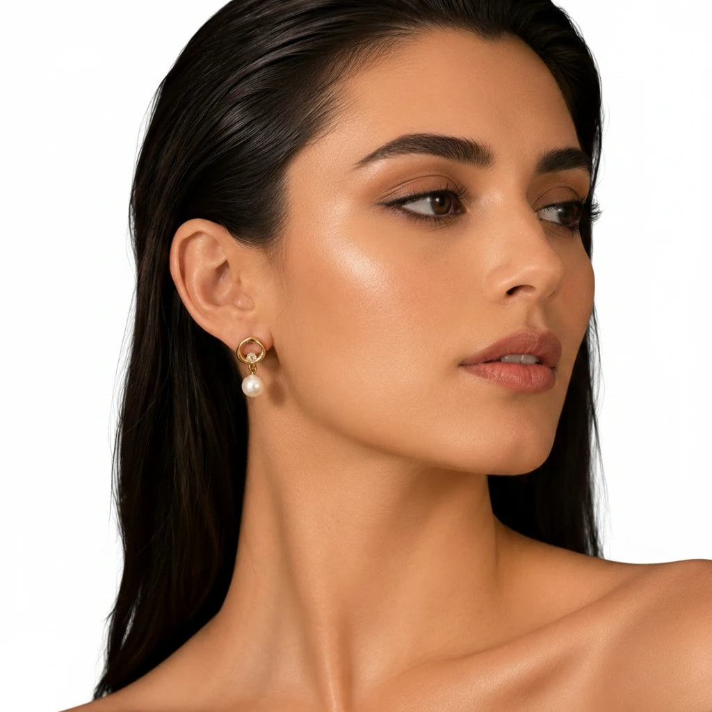 Elegant woman wearing EVAH JEWELS gold-tone circular pearl drop earrings featuring minimalist open-circle studs, sparkling crystal accents and delicate white pearl drops, styled with sleek dark hair against a clean white background.