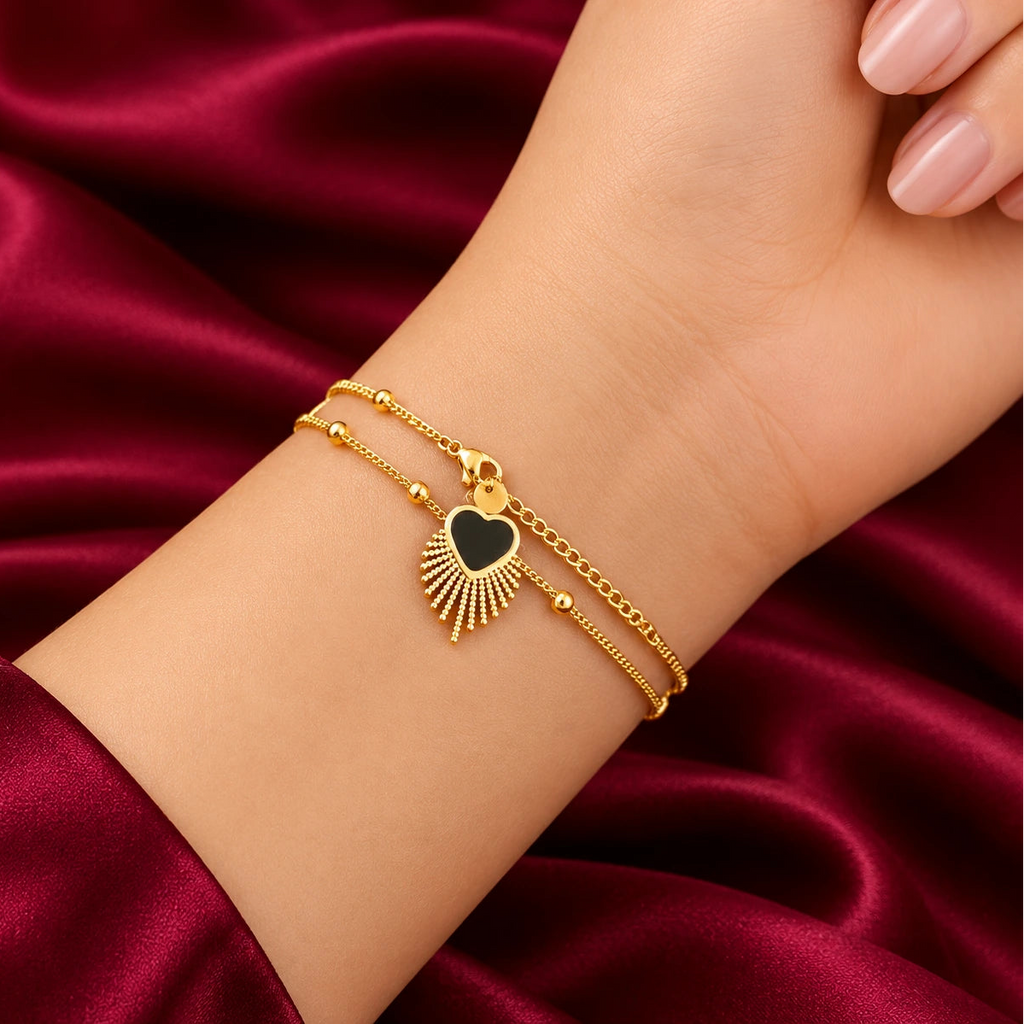 Discover effortless elegance with the EVAH JEWELS Gold Tone Black Heart Charm Bracelet for Women, a beautifully designed fashion jewellery accessory featuring a delicate gold-tone chain, polished golden bead accents and a striking black heart-shaped charm surrounded by an intricate sunburst-inspired fringe design. Elegantly showcased on a model's wrist against a luxurious burgundy satin background, this stylish bracelet highlights its sophisticated black and gold colour combination, graceful chain detailing