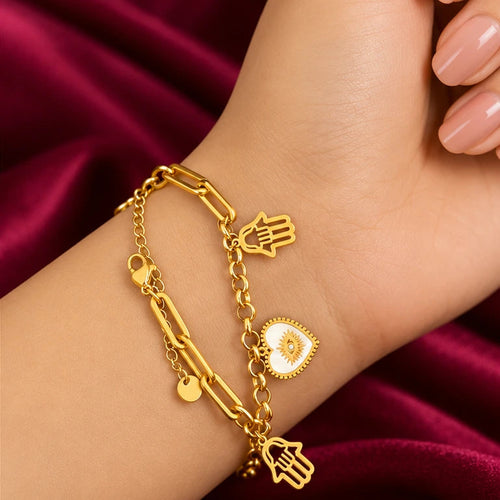 Elevate your everyday style with the EVAH JEWELS Gold Tone Hamsa Hand Charm Bracelet for Women, a beautifully designed fashion jewellery accessory featuring stylish gold-tone chain links, delicate Hamsa hand charms and an elegant white heart-shaped evil eye-inspired charm. Showcased gracefully on a model's wrist against a luxurious burgundy satin background, this trendy bracelet highlights its polished gold-tone finish, intricate charm detailing and fashionable layered chain design. The Hamsa hand and evil 