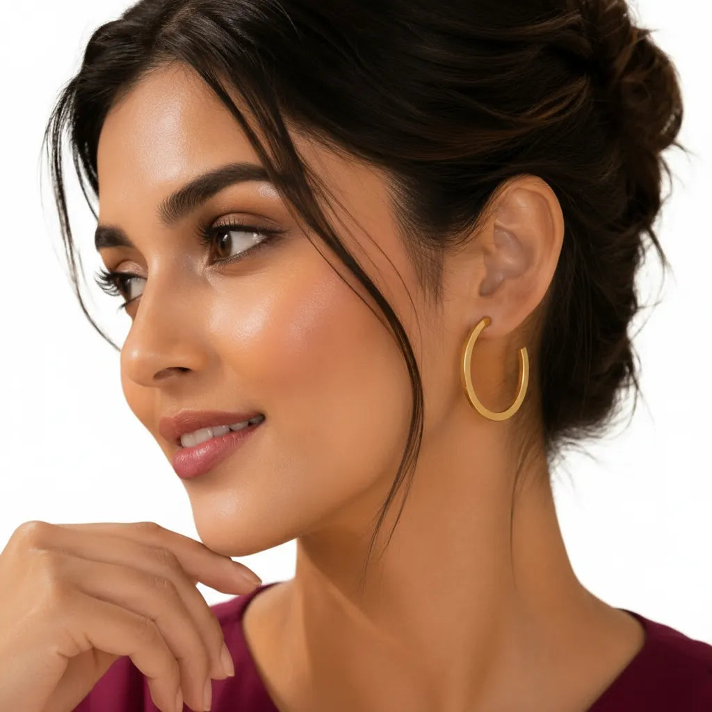 Woman wearing EVAH JEWELS minimalist gold-tone oval hoop earrings with a sleek polished open-hoop design, styled with an elegant burgundy outfit against a white background.