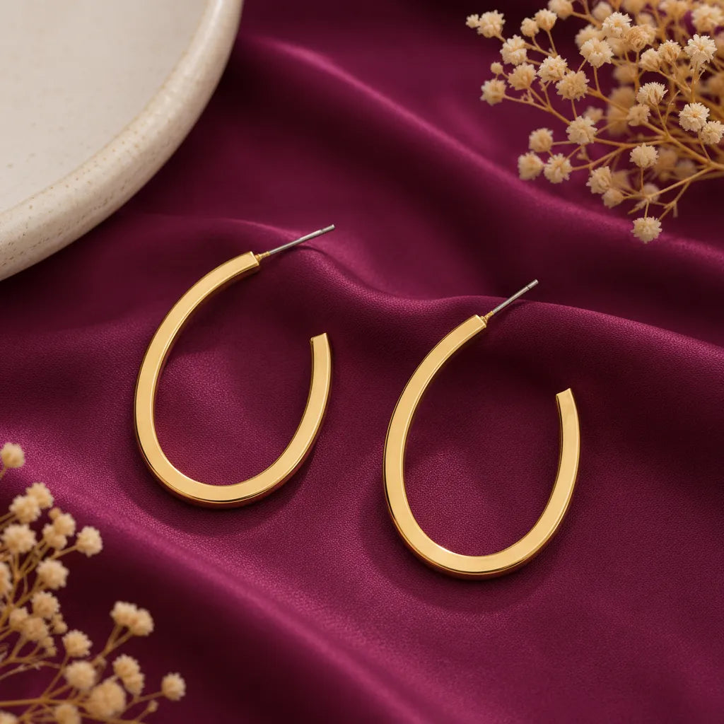 minimalist gold-tone oval hoop earrings for women featuring a sleek open-hoop design and polished finish, displayed on burgundy satin with delicate dried flowers by Evah Jewels.
