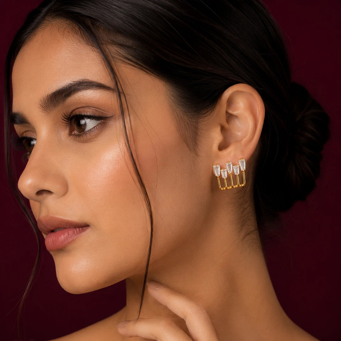 Elegant woman wearing EVAH JEWELS gold-tone geometric baguette crystal stud earrings featuring five sparkling rectangular clear stones arranged in a modern open-frame design, styled with a sleek low bun against a burgundy background.