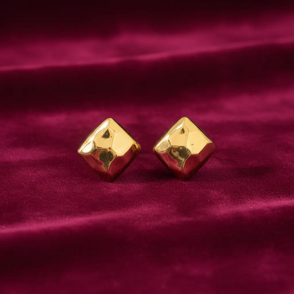 gold-tone geometric diamond-shaped stud earrings for women featuring a polished faceted surface, bold three-dimensional design and minimalist metallic finish, displayed on luxurious burgundy velvet fabric by Evah Jewels.