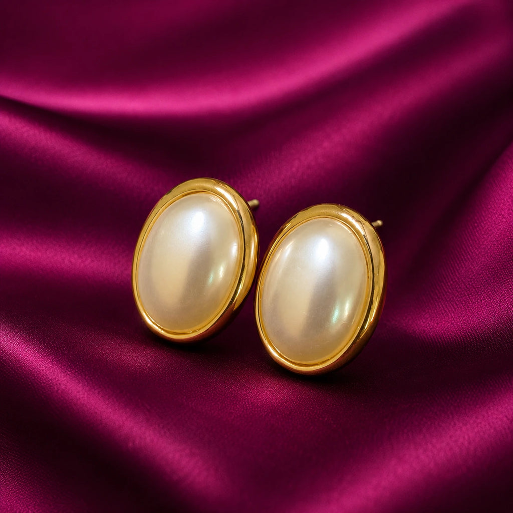 gold-plated oval pearl stud earrings for women featuring lustrous white pearl centres, polished gold-tone borders and a classic minimalist design, displayed on luxurious burgundy satin fabric by Evah Jewels.
