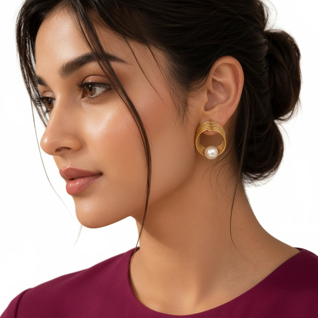 Elegant woman wearing EVAH JEWELS gold-tone spiral pearl statement stud earrings featuring layered concentric circular loops, a luminous white pearl accent and modern geometric styling, paired with a burgundy outfit and sophisticated low-bun hairstyle.