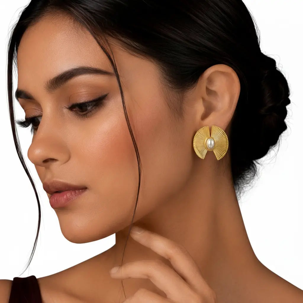 Elegant woman wearing EVAH JEWELS gold-tone sunburst pearl statement stud earrings featuring textured fan-shaped geometric detailing, oval white pearl centres and a radiant gold finish, styled with a sleek low bun against a white background.