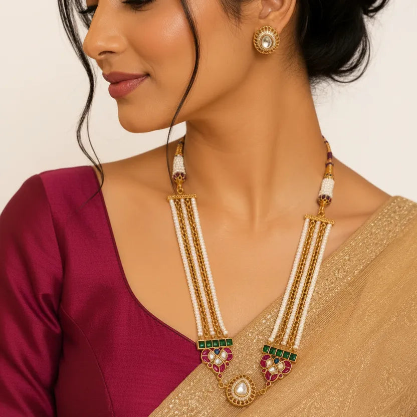 EVAH JEWELS traditional gold-tone Kundan pearl necklace set for women, featuring multiple white pearl bead strands, gold chains, green stone accents, pink floral detailing and a white stone pendant with matching round Kundan stud earrings. Model wearing elegant Indian ethnic jewellery with a maroon blouse and golden saree, ideal for weddings, festive celebrations and traditional occasions.